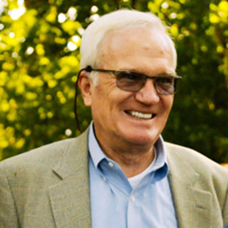 The image shows an older man with white hair and glasses. He is wearing a light-colored blazer over a blue shirt. He is smiling, and the background is blurred with green foliage. The man appears to be outdoors, possibly in a park or garden.